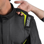 Close-up of a racing driver wearing a Sparco racing suit with black and grey fabric and yellow accents, showing the shoulder area.