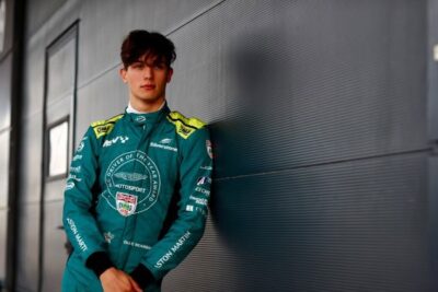 Young race car driver in Grand Prix Racewear suit standing against a modern building wall, ready for racing.