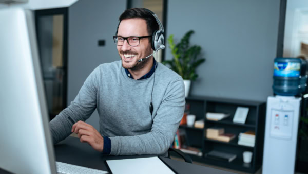 Customer support representative wearing a headset smiling at a computer in an office setting.