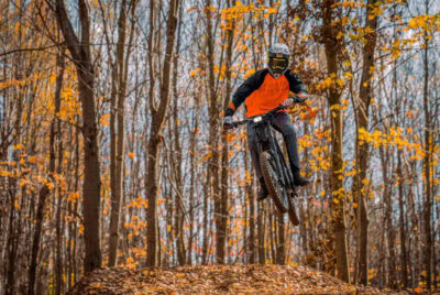 Mountain biker in orange and black gear jumps over a trail obstacle in a forest with autumn leaves.