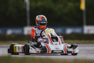 Go-kart racing driver in full gear on track, wearing a helmet with orange and blue accents, racing a white and red kart.