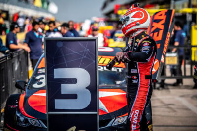 Race car driver in Grand Prix Racewear suit and helmet standing next to a race car with a large number 3 sign at a motorsport event.