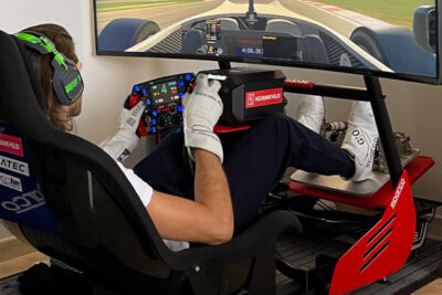 Race car driver in a simulator wearing headphones and gloves, focusing on the steering wheel and dashboard.