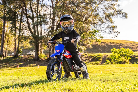 Young motocross rider in full gear on a blue dirt bike in a grassy field with trees in the background.
