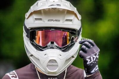 Close-up of a motocross rider wearing a white helmet, goggles with pink lenses, and black gloves, outdoors with green foliage in the background.