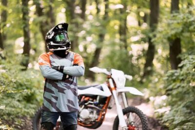 Motocross rider in full gear standing in a forest with a dirt bike in the background.