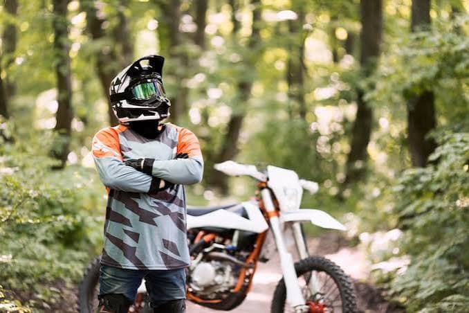 Motocross rider in full gear standing in a forest with a dirt bike in the background.