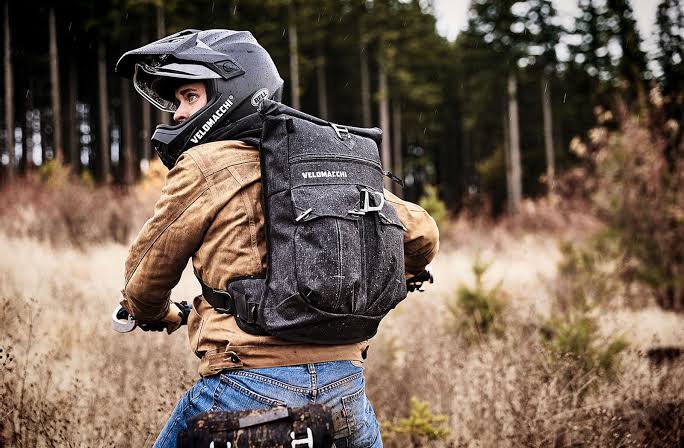Motorcycle rider wearing a helmet and protective gear with a backpack in a forest setting.
