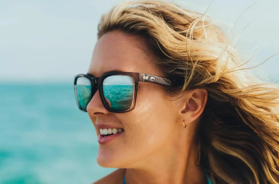 Close-up of a smiling woman wearing sunglasses at the beach, showcasing Grand Prix Racewear's stylish racing apparel.