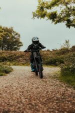 Child riding a mountain bike on a gravel trail surrounded by trees and greenery during daytime.