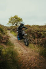 Child riding a mountain bike on a narrow dirt trail through a natural landscape with bushes and a tree in the background.
