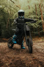 Child riding a black electric dirt bike in a forest, wearing a helmet and protective gear, on a dirt trail.